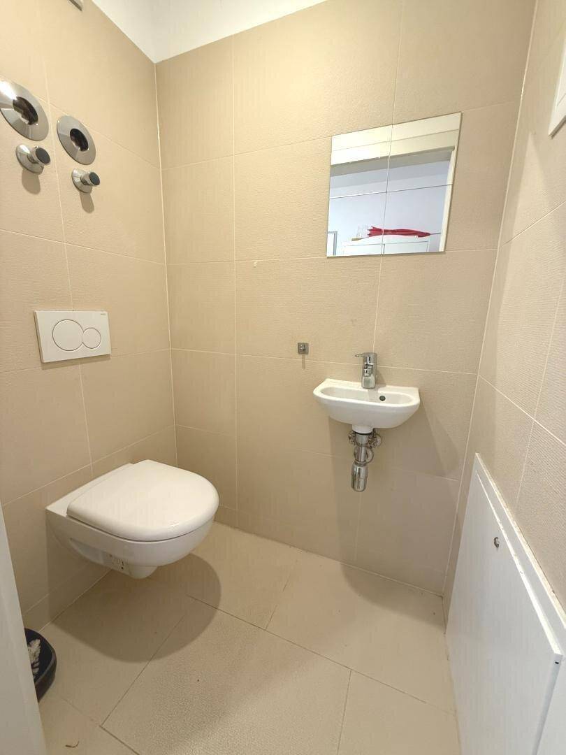 Separates WC mit Wandtoilette, kleinem Waschbecken und beigefarbenen Fliesen.