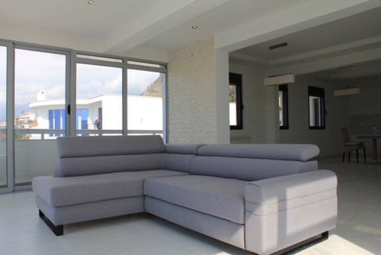 Helles Wohnzimmer mit Ecksofa und großen Fenstern, die viel Tageslicht hereinlassen.