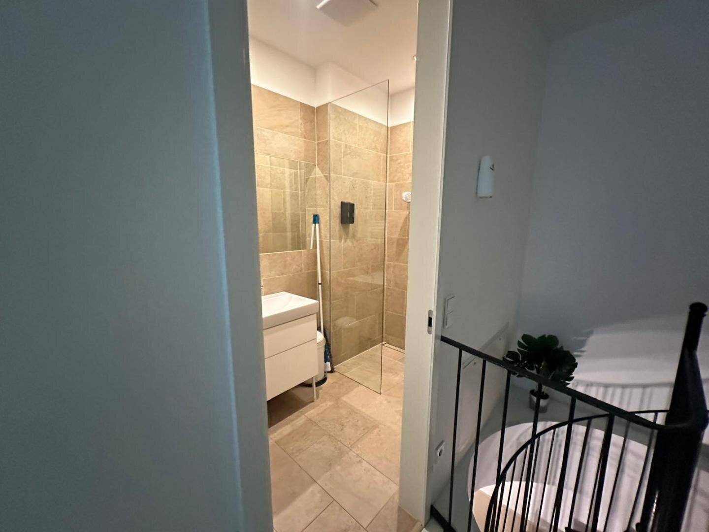 Modernes Badezimmer mit begehbarer Dusche und Einzelwaschbecken, sichtbar durch einen Türrahmen.