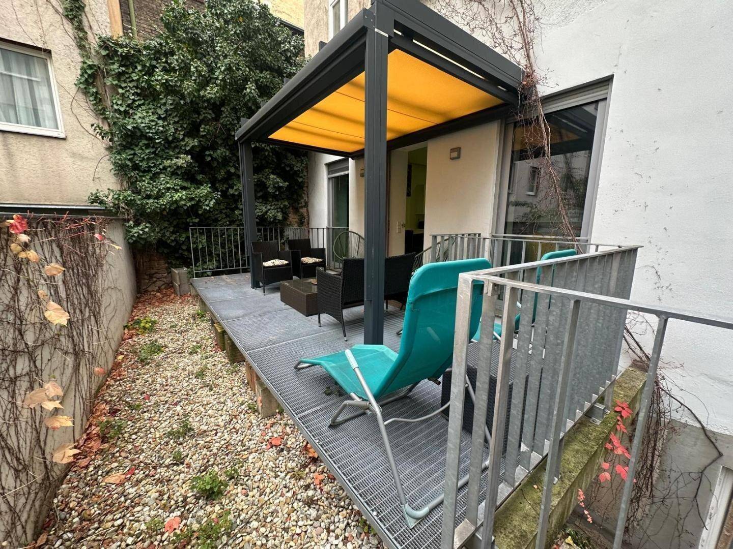 Außenterrasse mit Pergola und Sitzgelegenheiten, umgeben von Kies und Pflanzen.