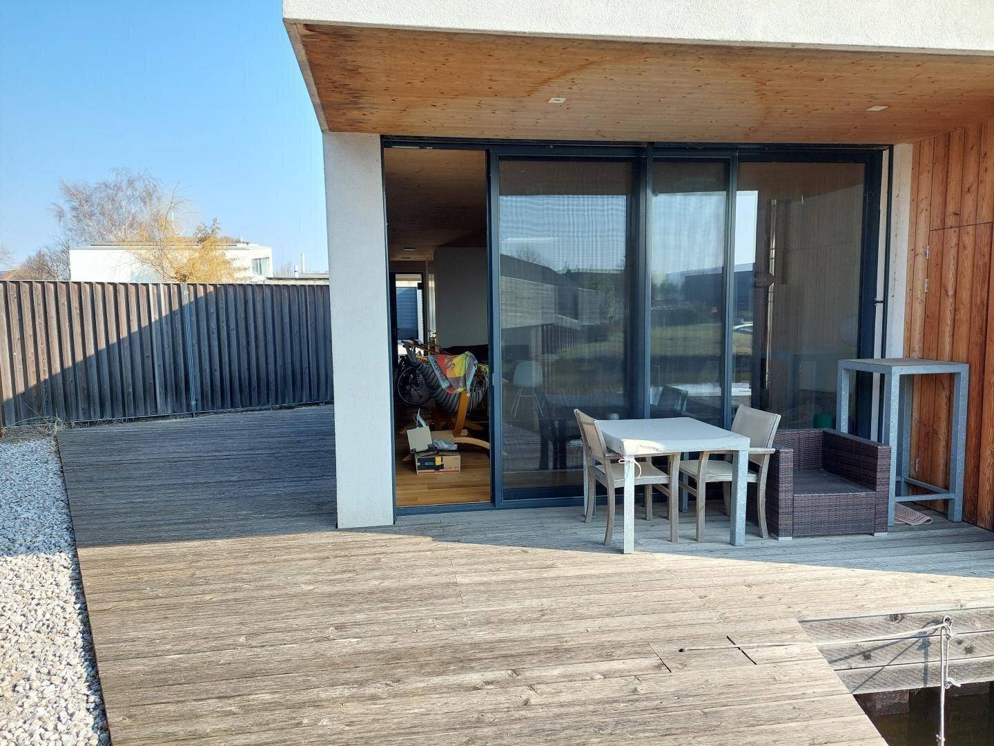 Moderne Holzterrasse mit Outdoor-Möbeln und großen Glasschiebetüren zum lichtdurchfluteten Innenbereich.