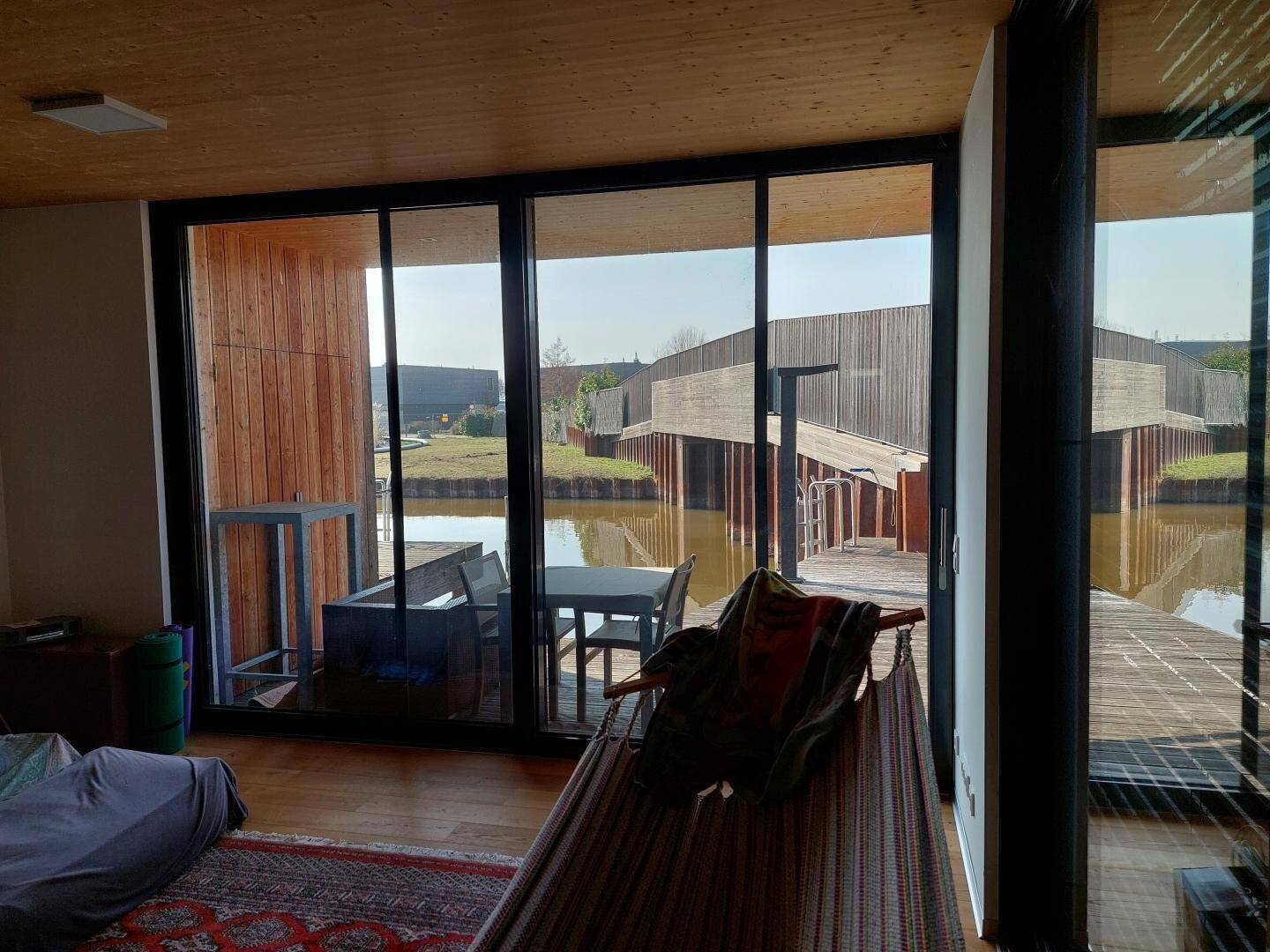 Heller Wohnbereich mit Hängematte, Holzbalkendecke und Panoramablick auf das Wasser und die Terrassen.