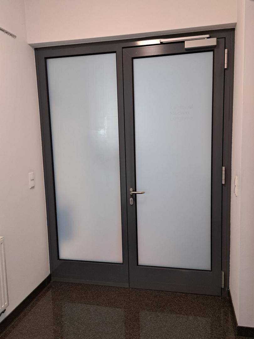 Eingangsbereich mit einer doppelten Glastür und dunklem, glänzendem Fliesenboden.