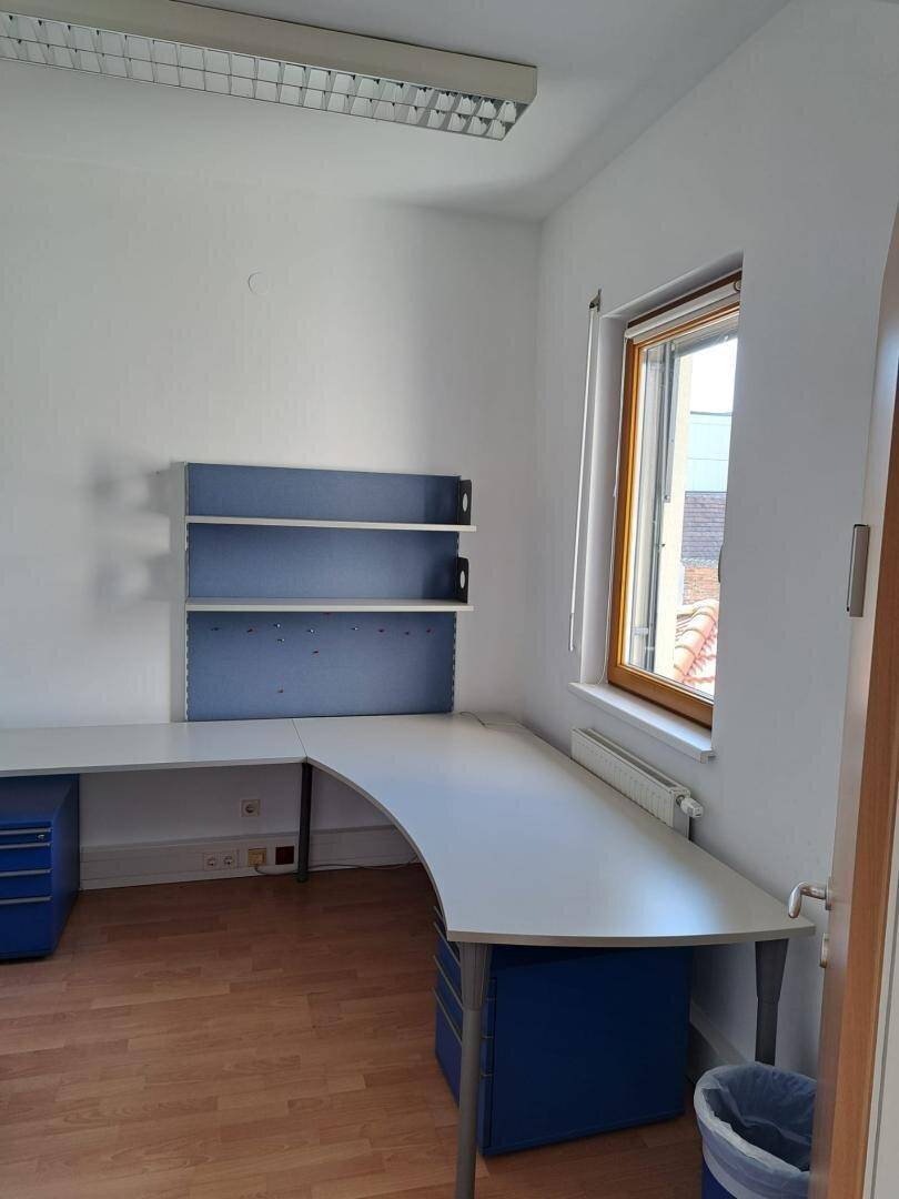 Eckbereich eines Büros mit Schreibtisch, blauen Regalen und einem Fenster mit Heizkörper.