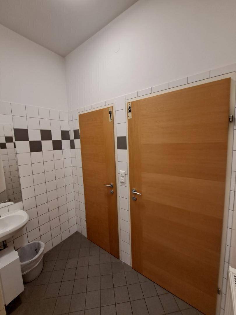 Toilettenbereich mit zwei Holztüren, weiß-grauen Fliesen und einem Waschbecken.