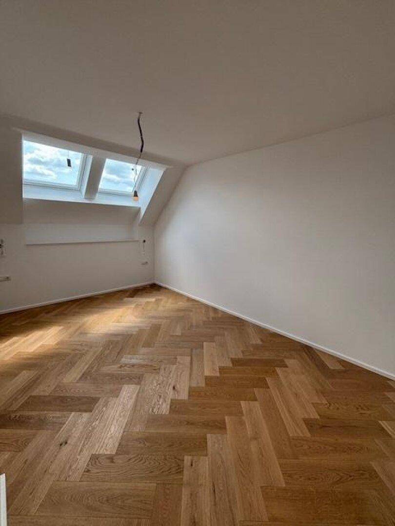 Geräumiges Dachzimmer mit Fischgrätparkett, weißen Wänden und viel Tageslicht.