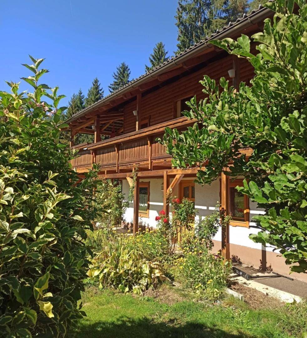 Traditionelles Holzhaus mit Balkon und üppigem Garten, umgeben von hohen Bäumen unter blauem Himmel.