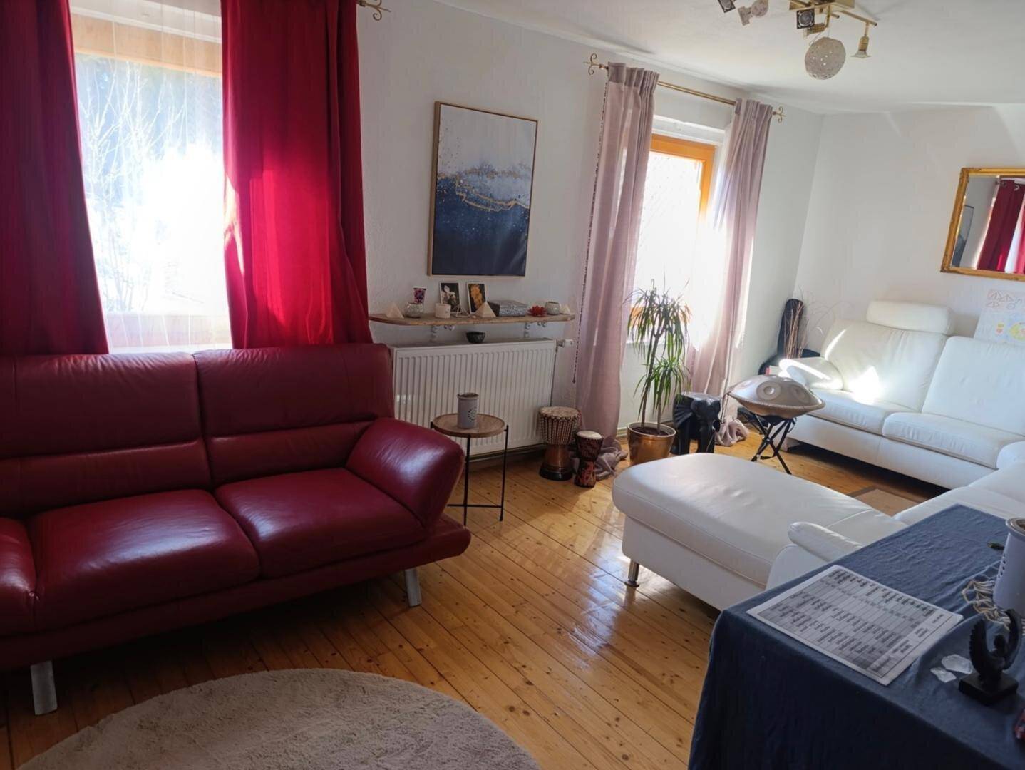 Helles Wohnzimmer mit Holzdielenboden, roten und weißen Sofas und großen Fenstern mit roten Vorhängen.