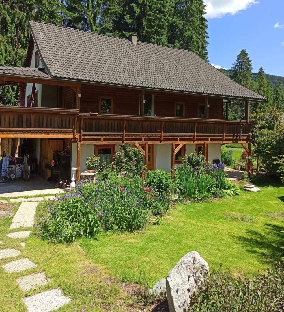 Charmantes Holzhaus mit Balkon und blühendem Garten, umgeben von hohen Nadelbäumen an einem sonnigen Tag.
