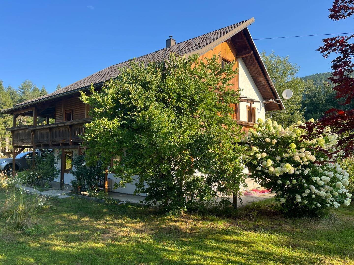 Haus mit Holz- und Putzfassade, umgeben von Bäumen und blühenden Sträuchern im Sonnenlicht.