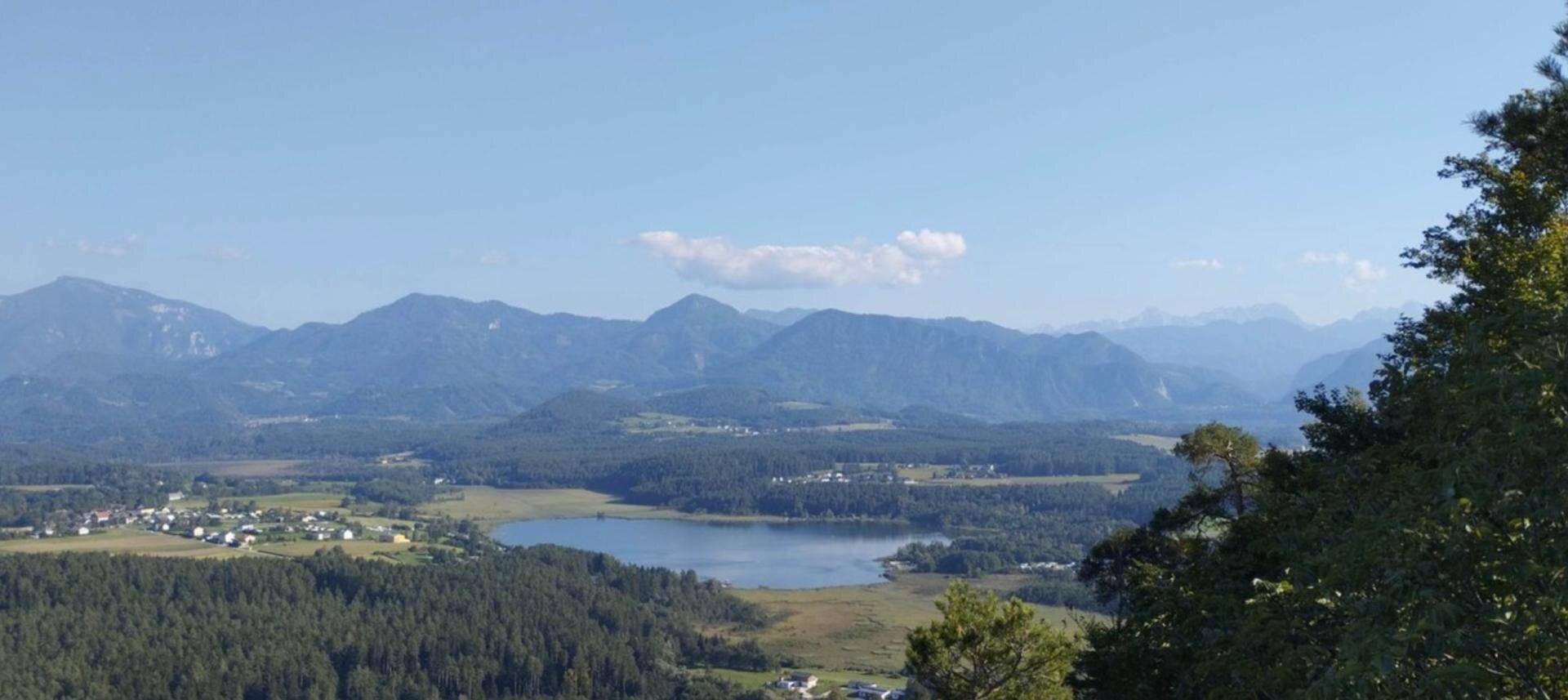 Panoramablick auf einen See, umgeben von Wäldern und Bergen unter blauem Himmel.