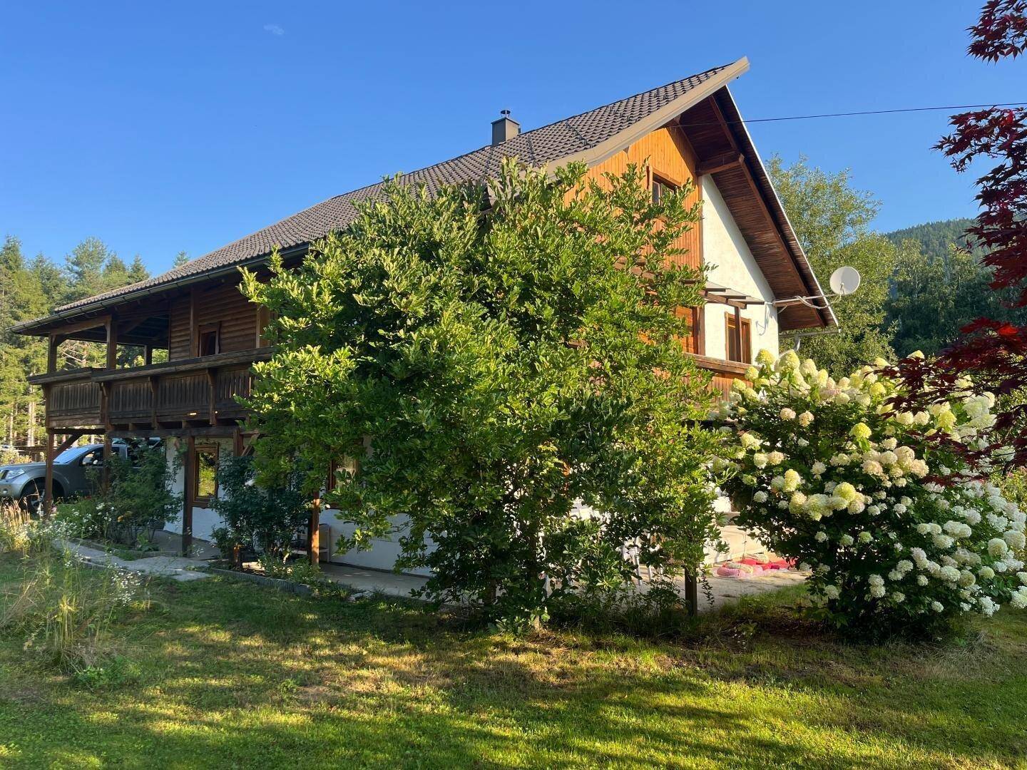 Haus mit Holz- und Putzfassade, umgeben von Bäumen und blühenden Sträuchern im Sonnenlicht.