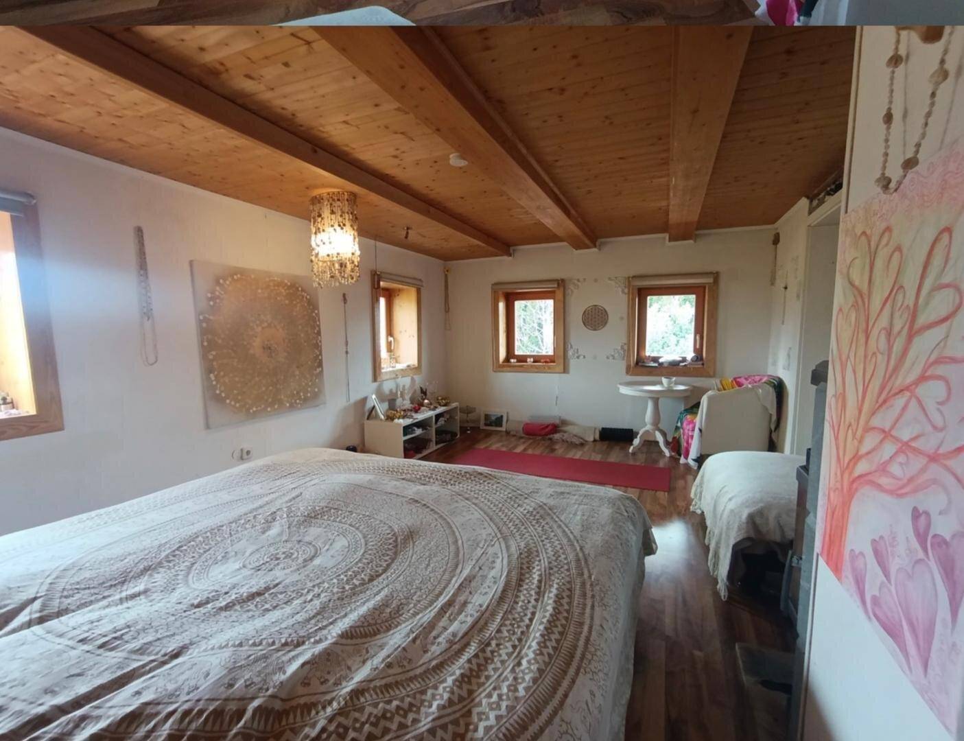 Helles Schlafzimmer mit Holzbalkendecke, großem Bett und mehreren Fenstern, die viel Licht hereinlassen.