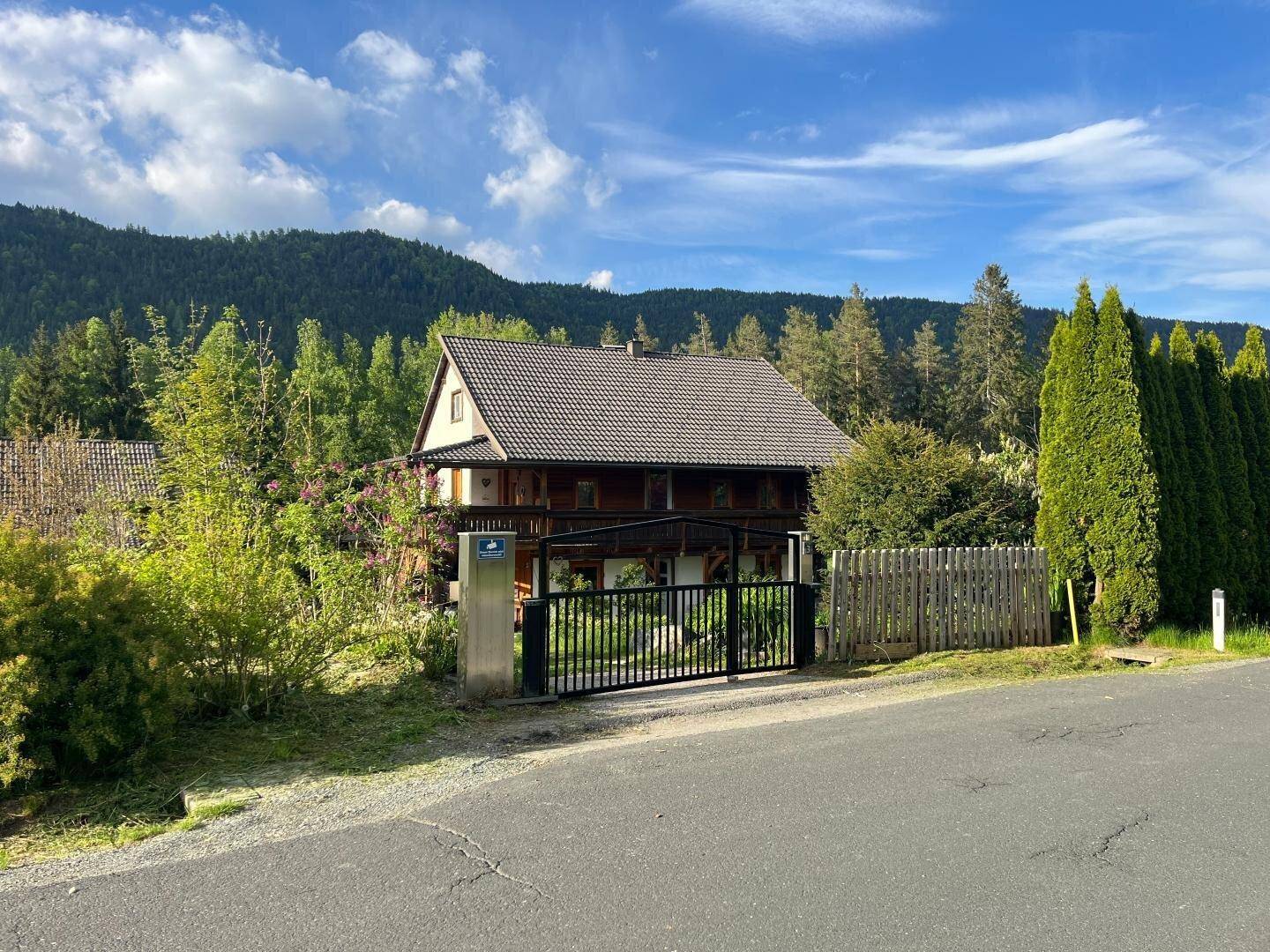 Traditionelles Haus mit Satteldach, umgeben von hohen Hecken und einem schwarzen Eingangstor an der Straße.