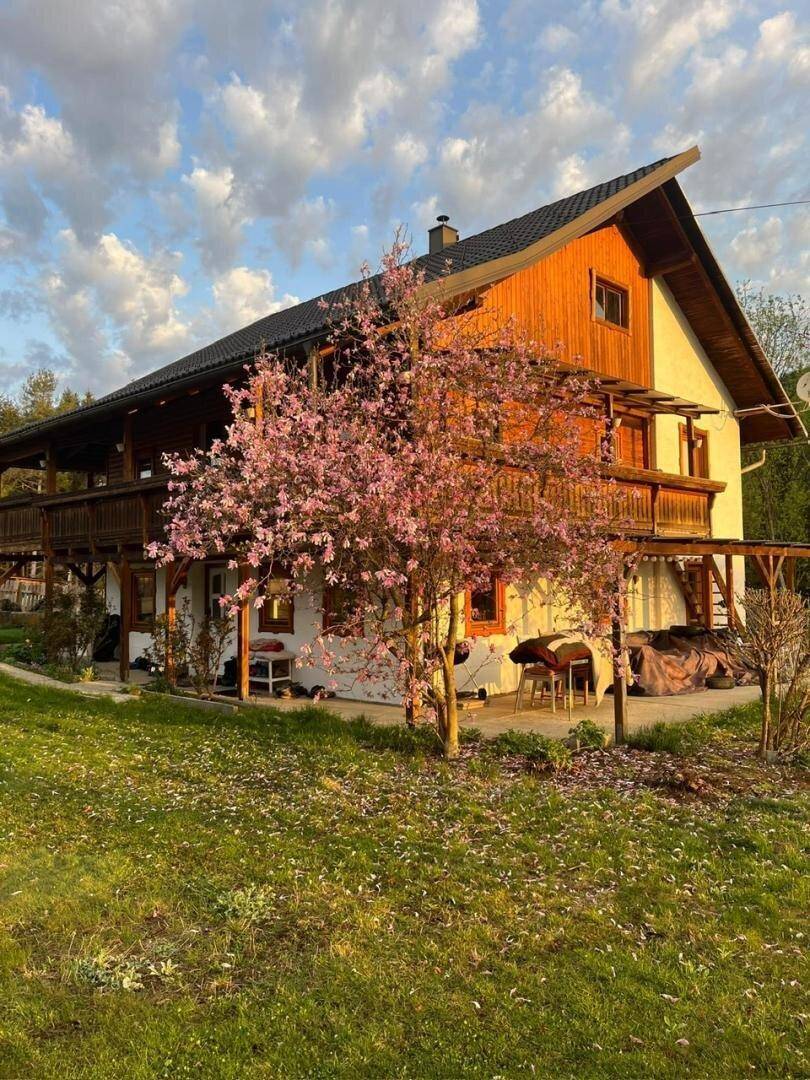 Charmantes Haus mit Holzverkleidung und Balkonen, umgeben von einem blühenden Baum und grüner Wiese.