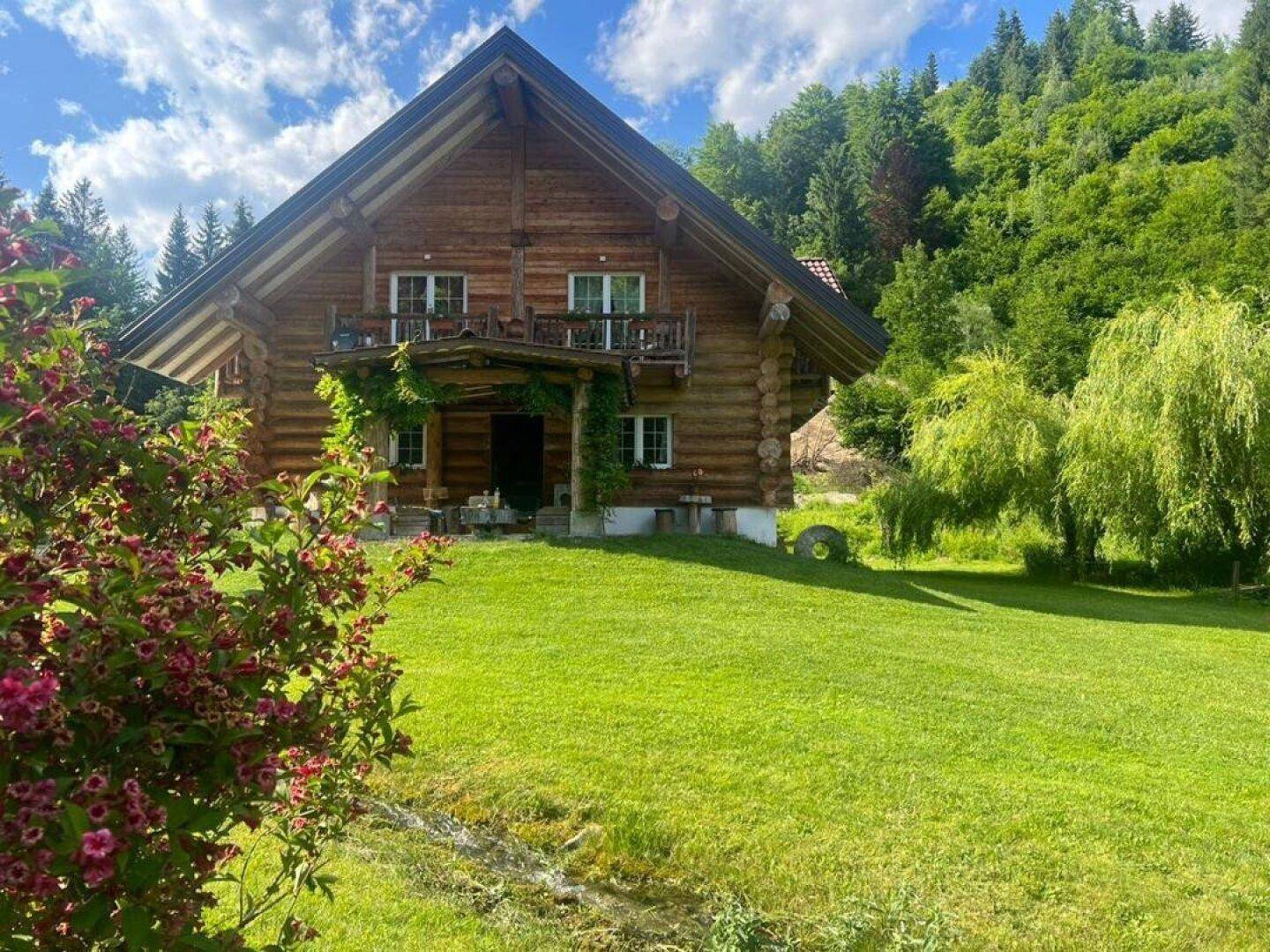 Idyllisches Blockhaus in weitläufiger grüner Landschaft mit Rasenfläche und dichtem Wald im Hintergrund.