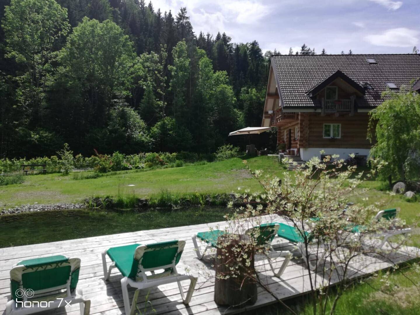 Holzterrasse mit Liegestühlen und Blick auf das Blockhaus und die grüne Hügellandschaft.
