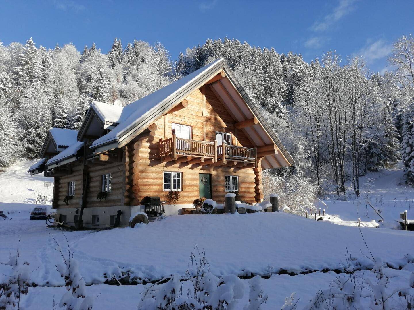 Winterliche Ansicht des Blockhauses, eingebettet in eine verschneite Landschaft mit Tannenbäumen.
