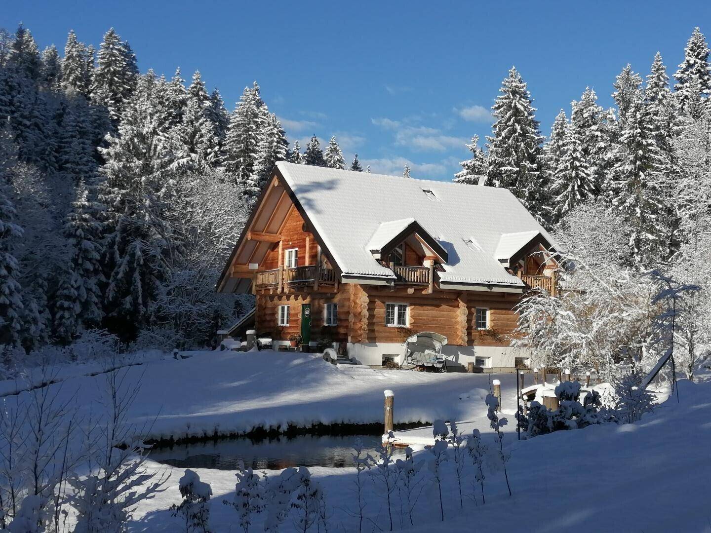 Blockhaus im Winter mit verschneiter Umgebung und einem teilweise zugefrorenen Teich.