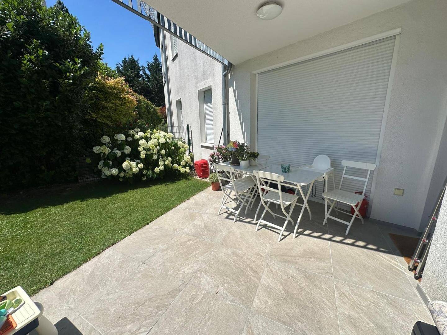 Sonnige Terrasse mit Gartenmöbeln, Rasenfläche und Außenjalousien.