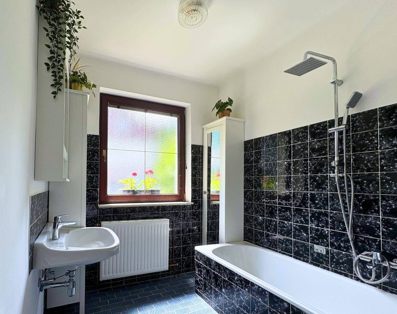 Badezimmer mit schwarz-weißen Fliesen, Badewanne mit Duschfunktion und Fenster.