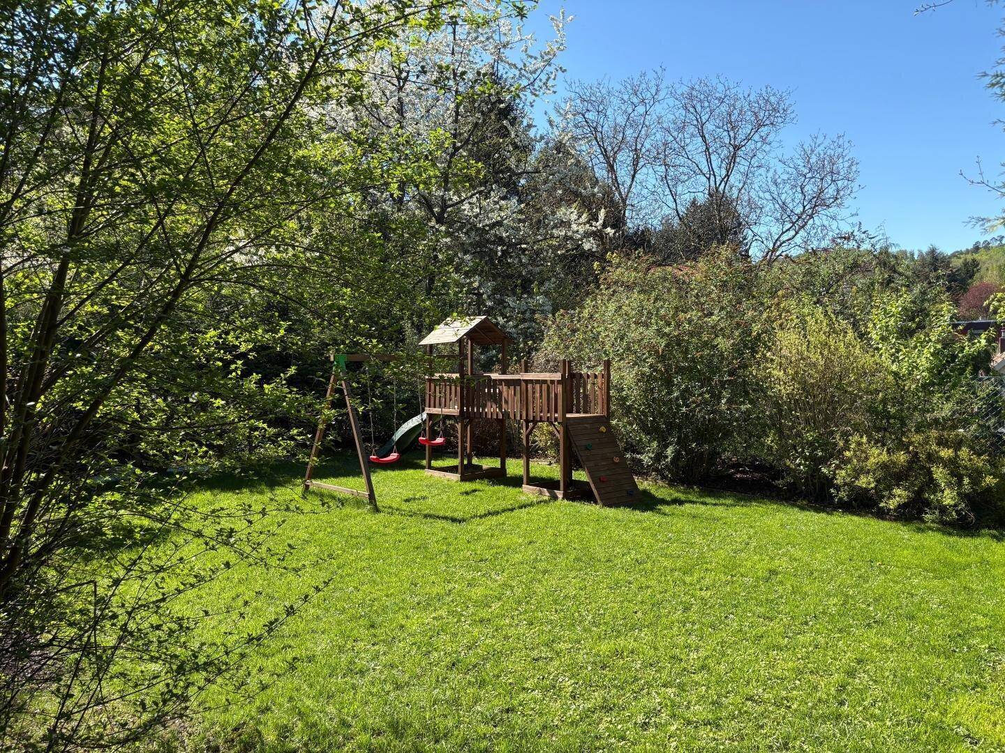 Großer Gartenbereich mit Holzspielplatz, Schaukeln und viel Grün.