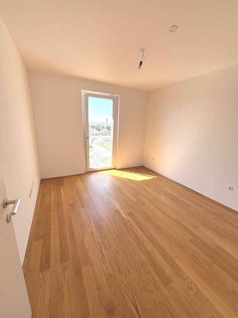 Leeres Zimmer mit Holzboden und großem Fenster, das viel Tageslicht hereinlässt.