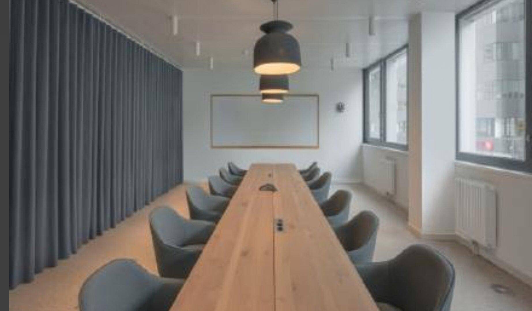 Langer Konferenztisch mit modernen Stühlen in einem hellen Raum mit großen Fenstern und eleganten Vorhängen.
