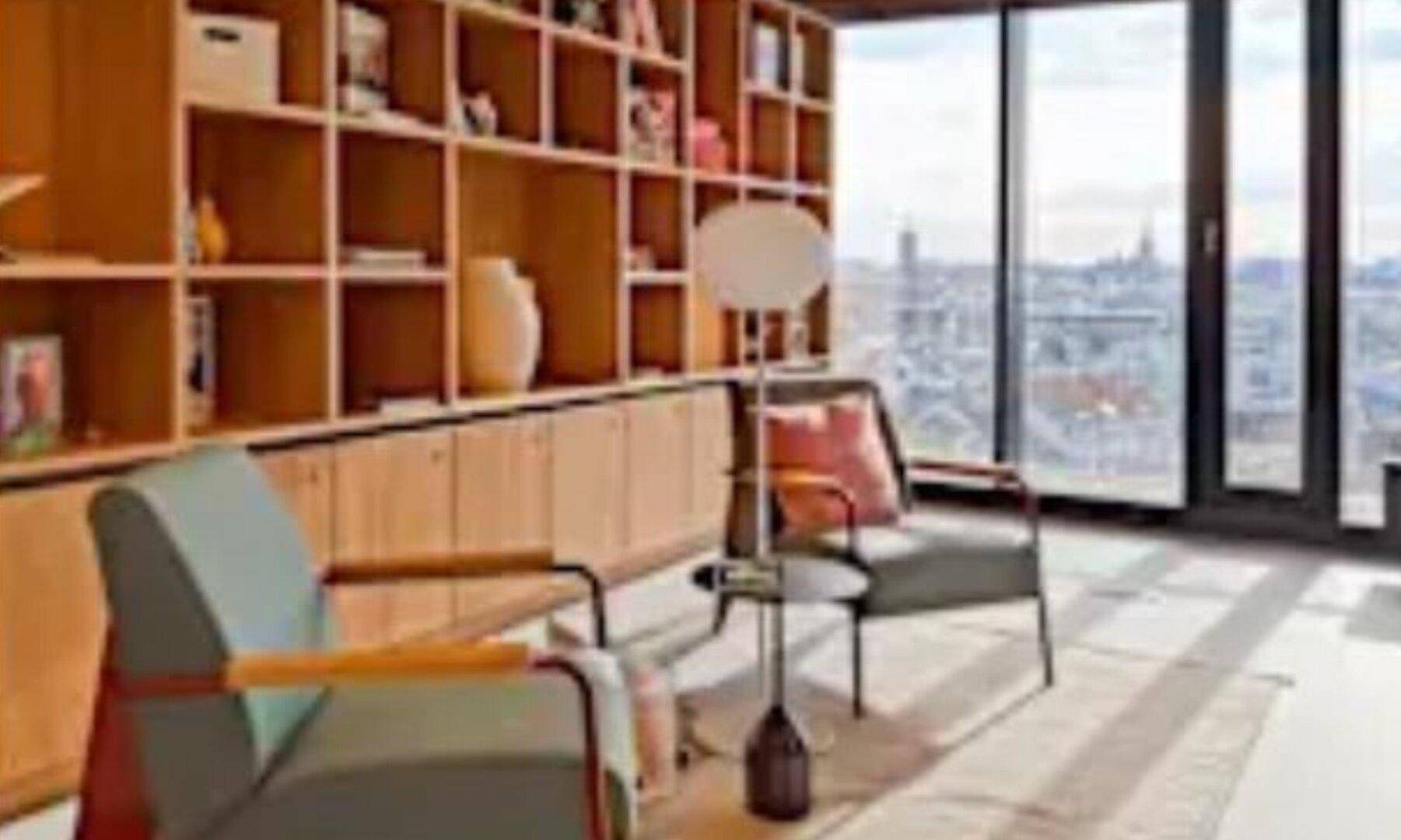 Heller Loungebereich mit Holzregalen, bequemen Sesseln und weitem Stadtblick durch große Fenster, perfekt zum Entspannen.