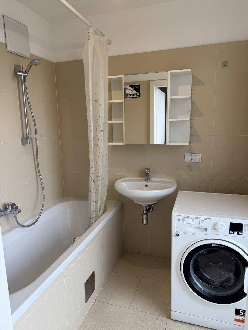 Helles Badezimmer mit Badewanne, Waschbecken und Waschmaschine, in beigen Fliesen gehalten.