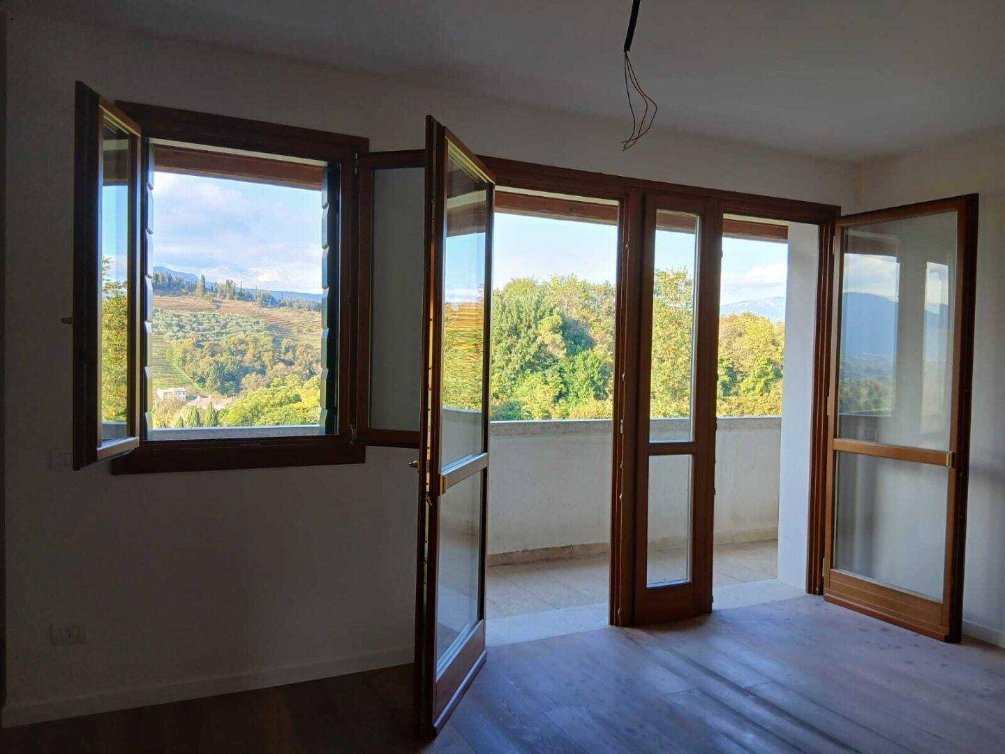 Heller Raum mit Holzboden und großen Fenstertüren, die zu einem Balkon mit Weitblick führen.