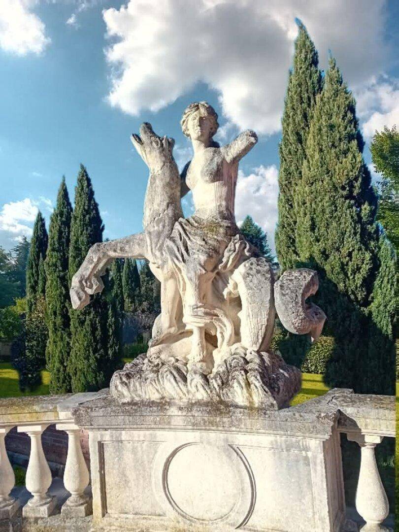 Antike Steinstatue auf einem Sockel, umgeben von hohen Zypressen und blauem Himmel.