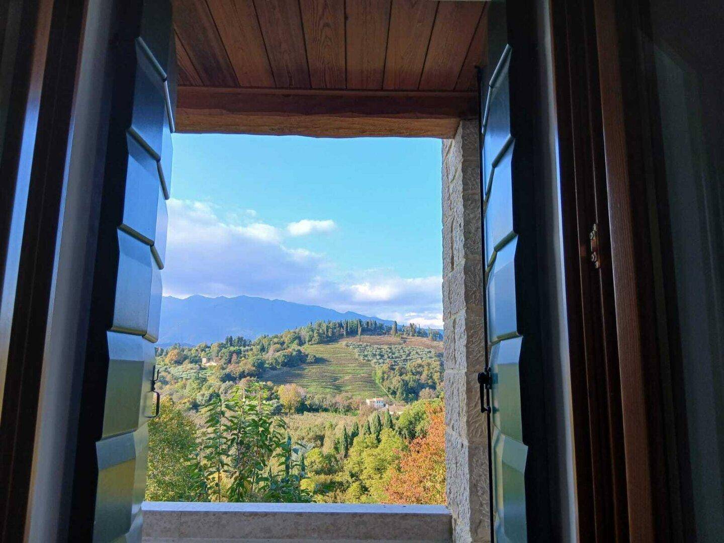 Blick aus einem Fenster mit geöffneten Fensterläden auf eine grüne Hügellandschaft und Berge.