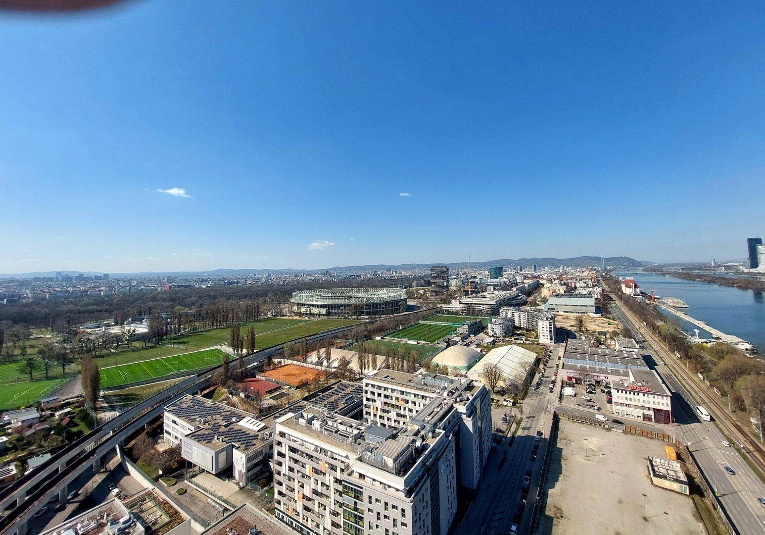 Weitläufiger Blick über die Stadt, den Fluss und das Stadion unter einem klaren blauen Himmel.