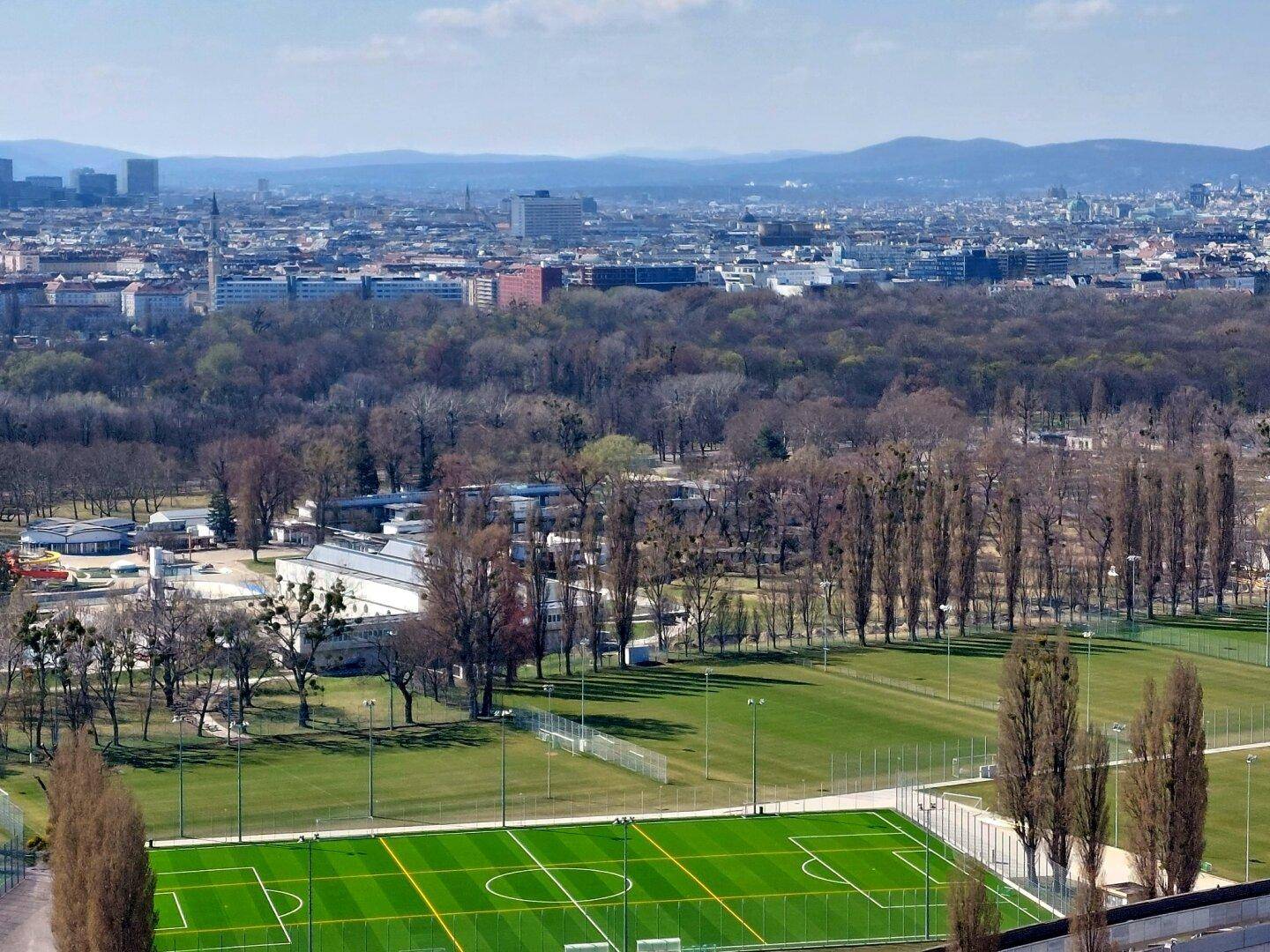 Weitläufige Grünflächen mit Sportfeldern und Bäumen, im Hintergrund die Stadt Wien.
