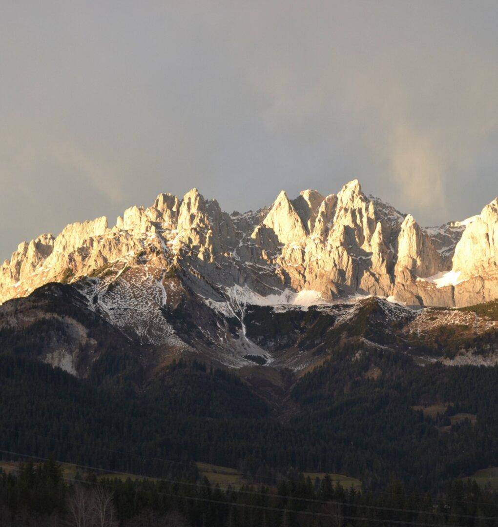 Majestätische Berggipfel im warmen Licht der Abendsonne mit schneebedeckten Hängen und dunklen Wäldern.