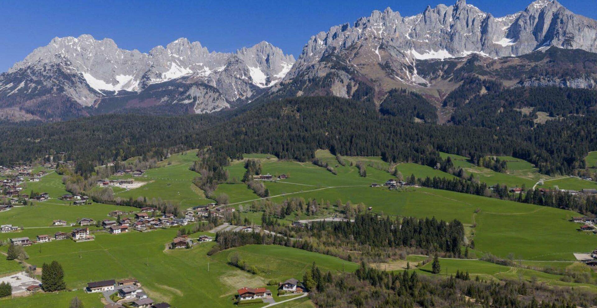 Panoramablick auf ein malerisches Dorf in einem grünen Tal, umgeben von beeindruckenden Bergketten und Wäldern.