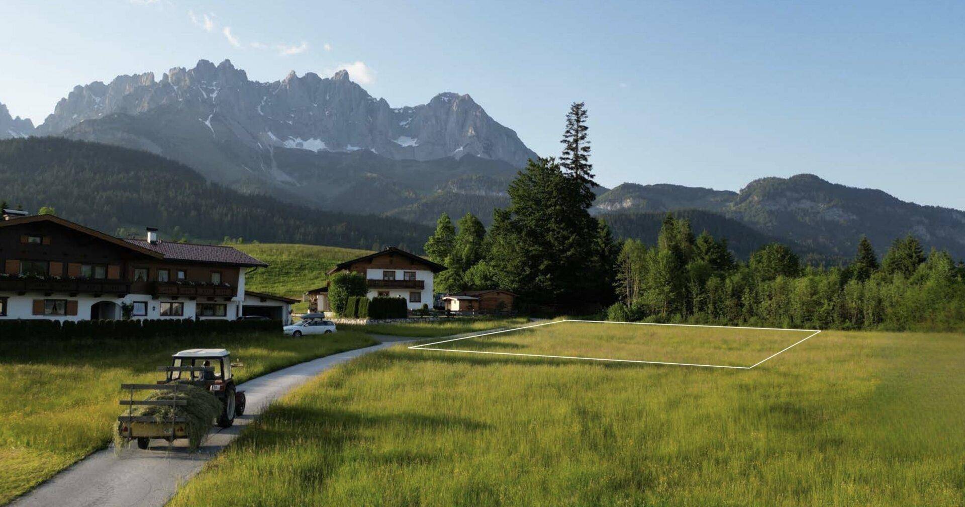 Grünes Baugrundstück in ländlicher Umgebung mit traditionellen Häusern, einem Traktor und majestätischem Bergblick.