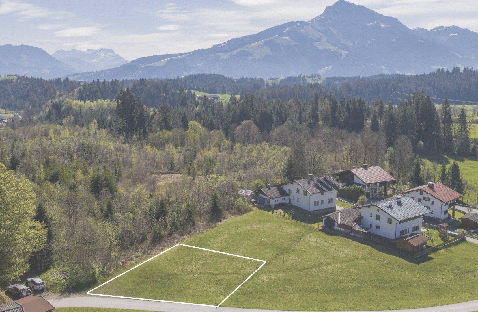 Luftaufnahme eines Baugrundstücks in ländlicher Umgebung mit bewaldeten Hügeln und Bergen.