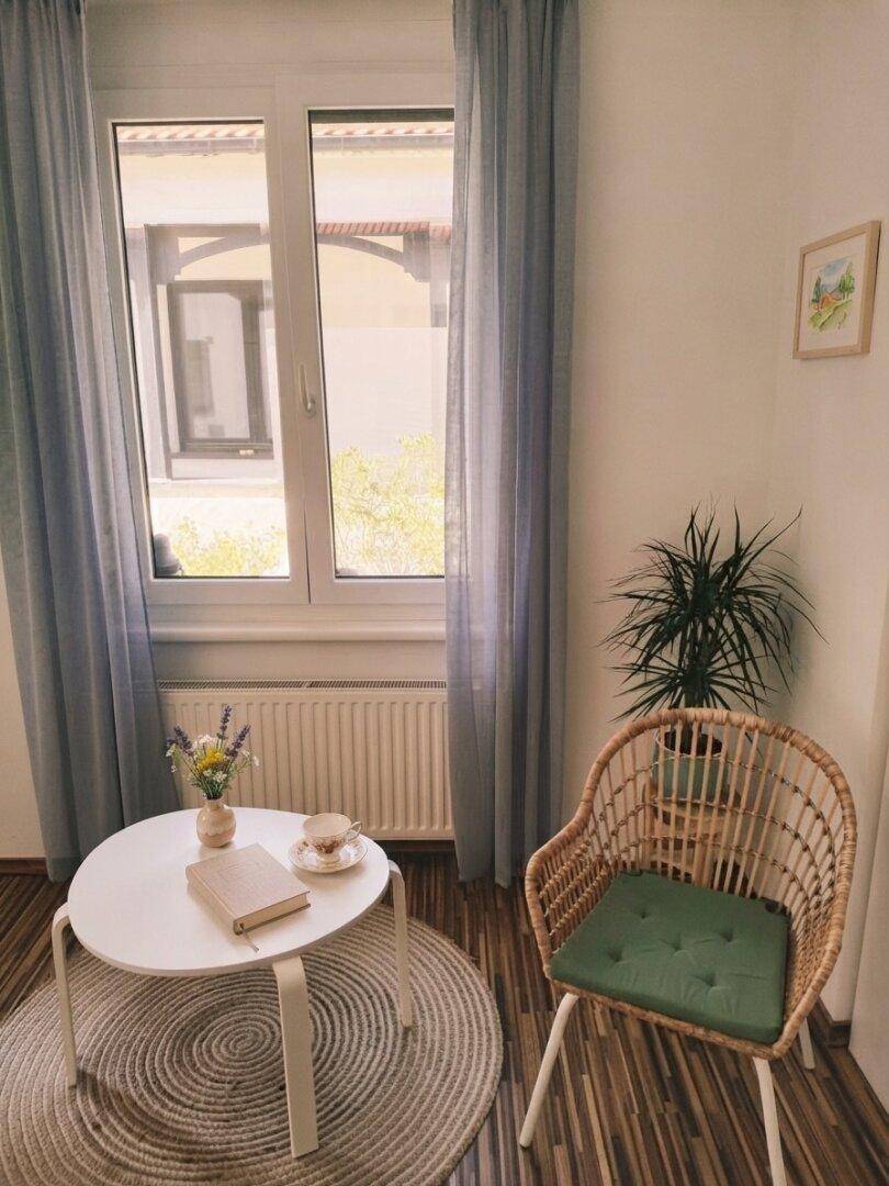 Gemütliche Sitzecke mit Rattan-Sessel, kleinem Tisch und Blick durch das Fenster ins Grüne.