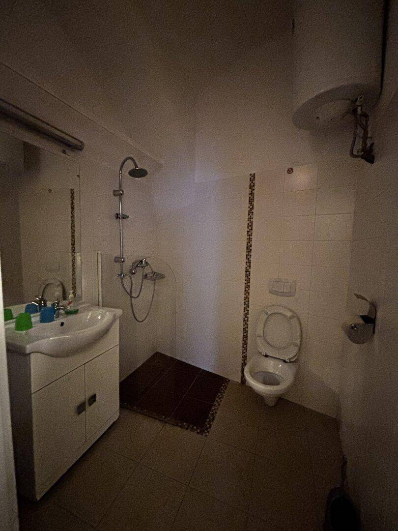 Badezimmer mit Dusche, WC, Waschbecken und Warmwasserboiler, schwach beleuchtet.