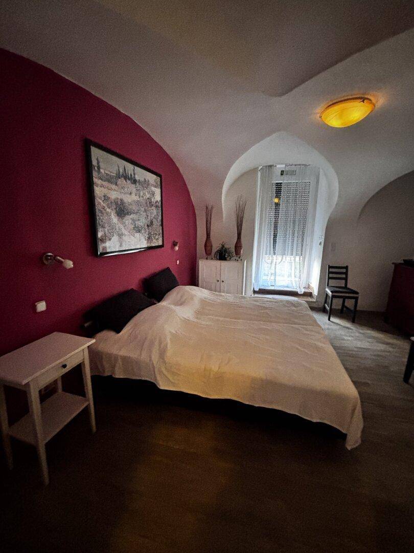 Schlafzimmer mit Doppelbett, roter Akzentwand und gewölbter Decke, Fenster mit Rollladen.
