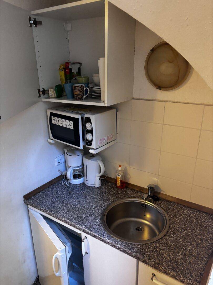 Kleine Küchenzeile mit Spüle, Mikrowelle, Kaffeemaschine und Wasserkocher, geflieste Rückwand.