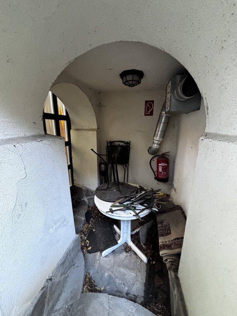 Gewölbter Durchgangsbereich mit einem Tisch und einem Feuerlöscher, der ins Freie führt.