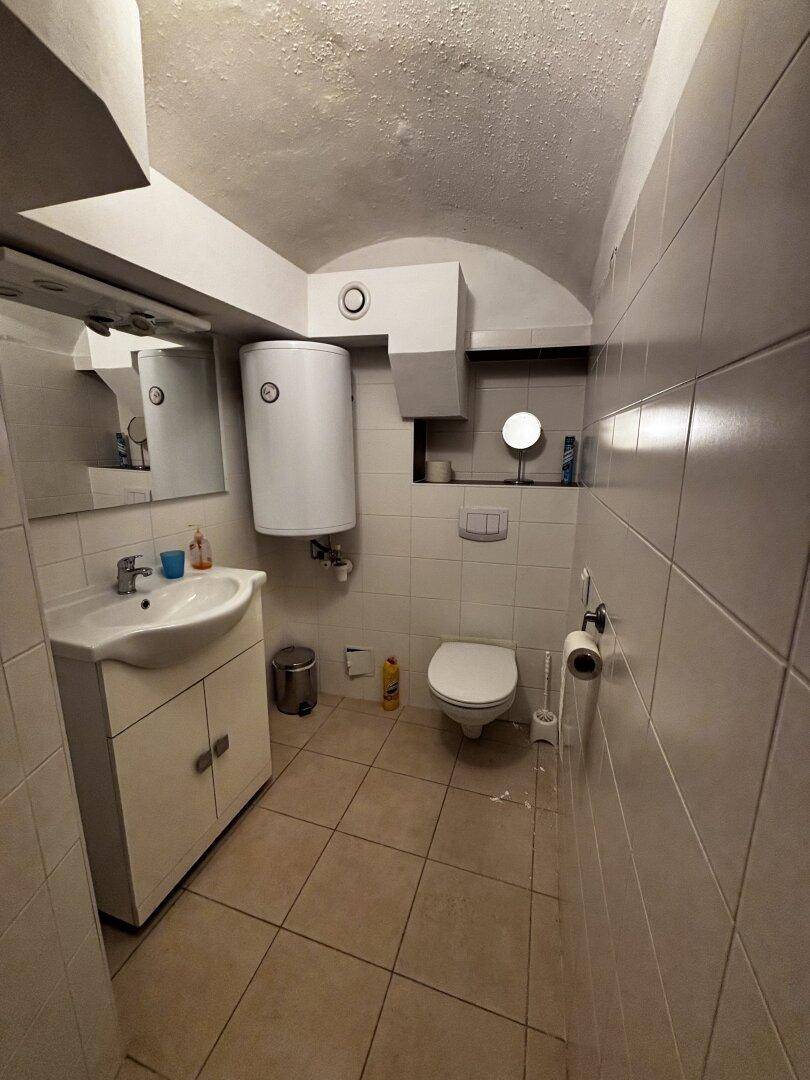 Einfaches Badezimmer mit Toilette, Waschbecken, Spiegel und Warmwasserbereiter in gefliestem Raum.