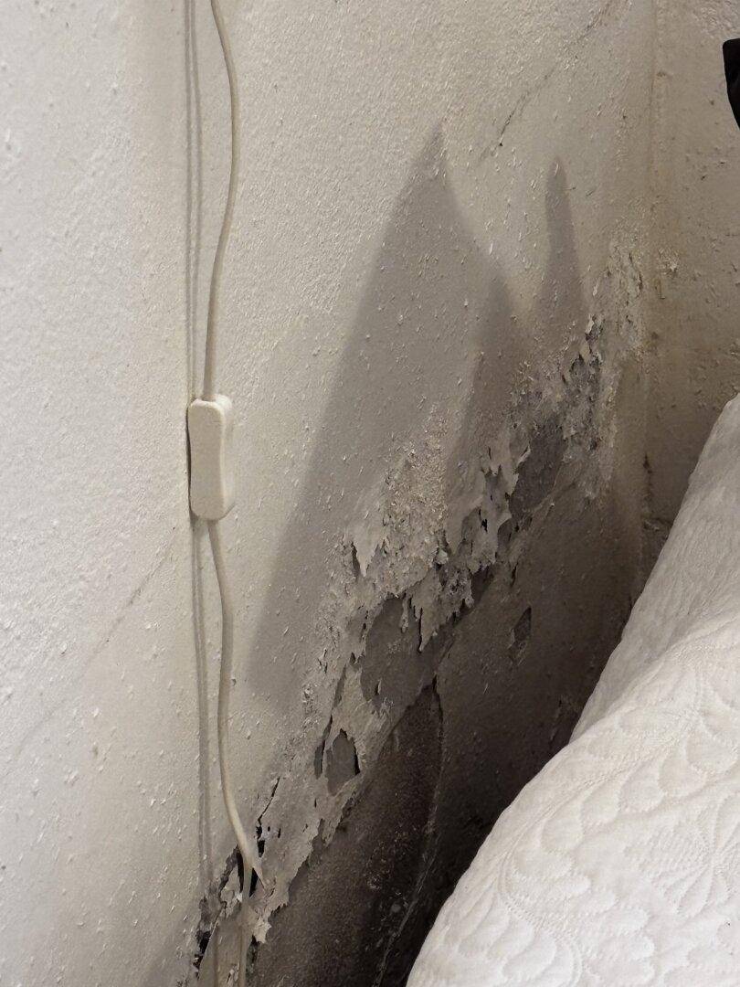 Nahaufnahme einer Wand neben einem Bett mit abblätternder Farbe und Feuchtigkeitsspuren.
