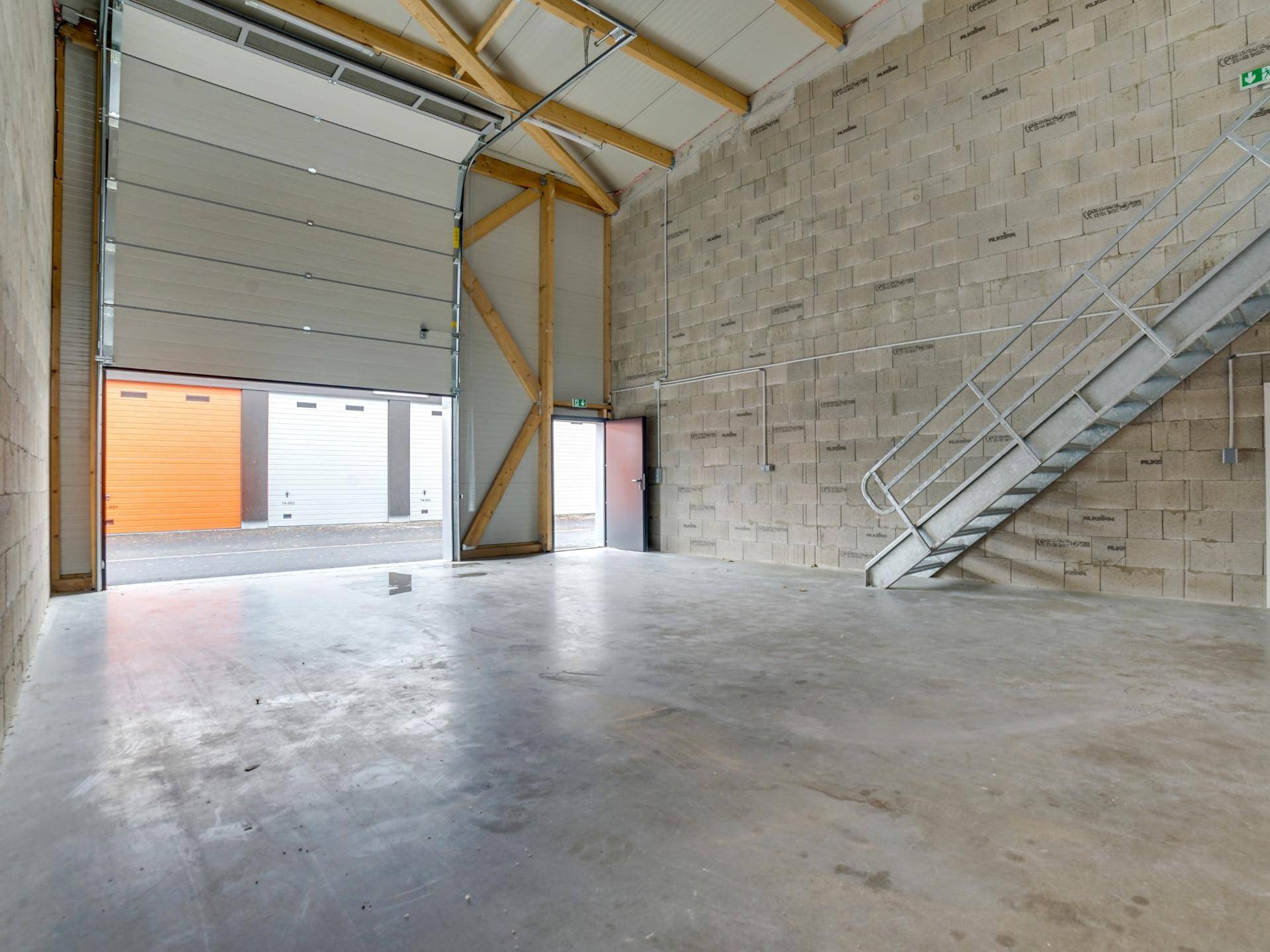 Großer, leerer Lagerraum mit Betonboden, Betonsteinwänden und einer Metalltreppe, Rolltor geöffnet.