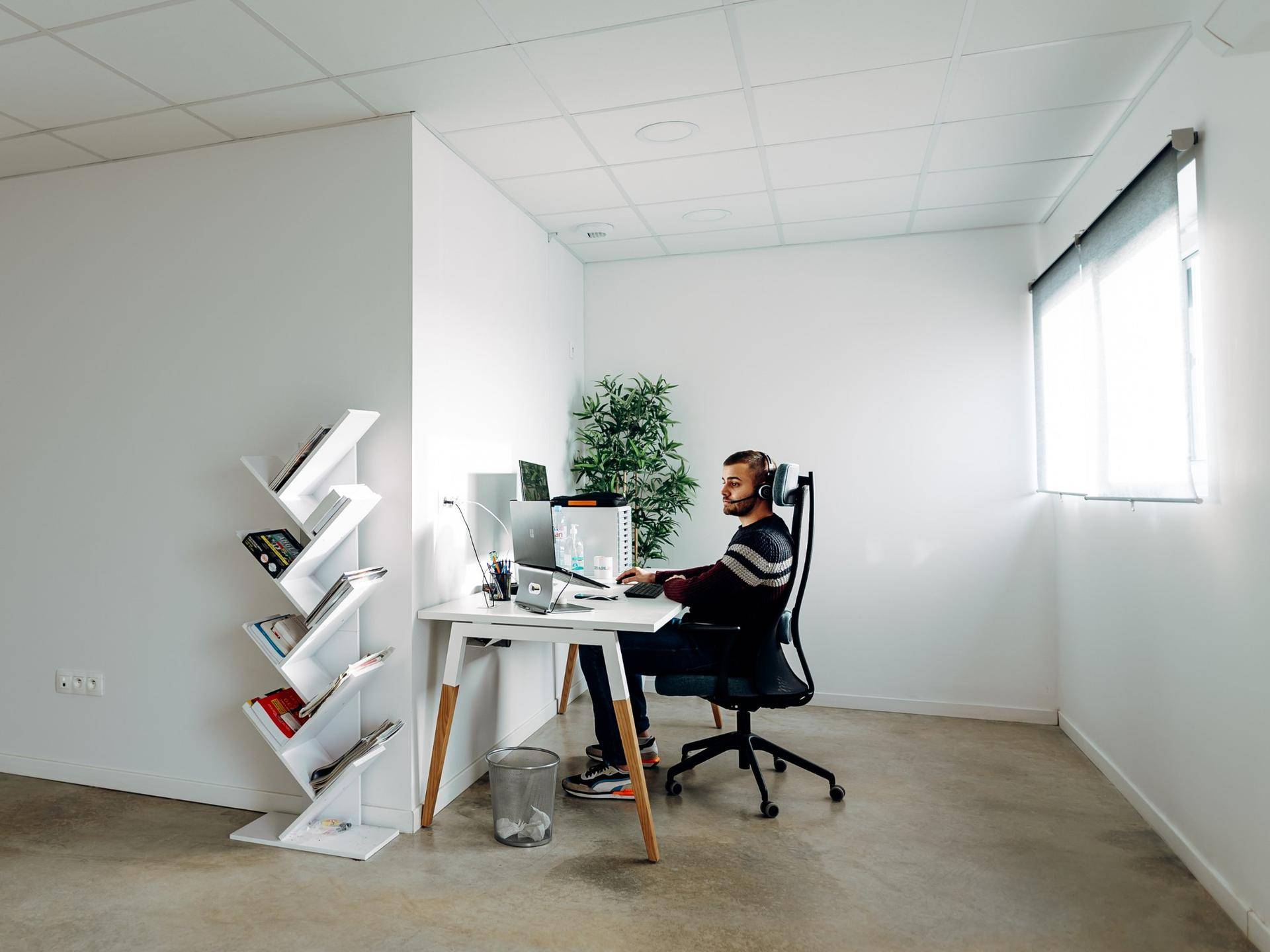 Moderner Bürobereich mit einer Person am Schreibtisch, weißen Wänden und einem Fenster mit Jalousie.