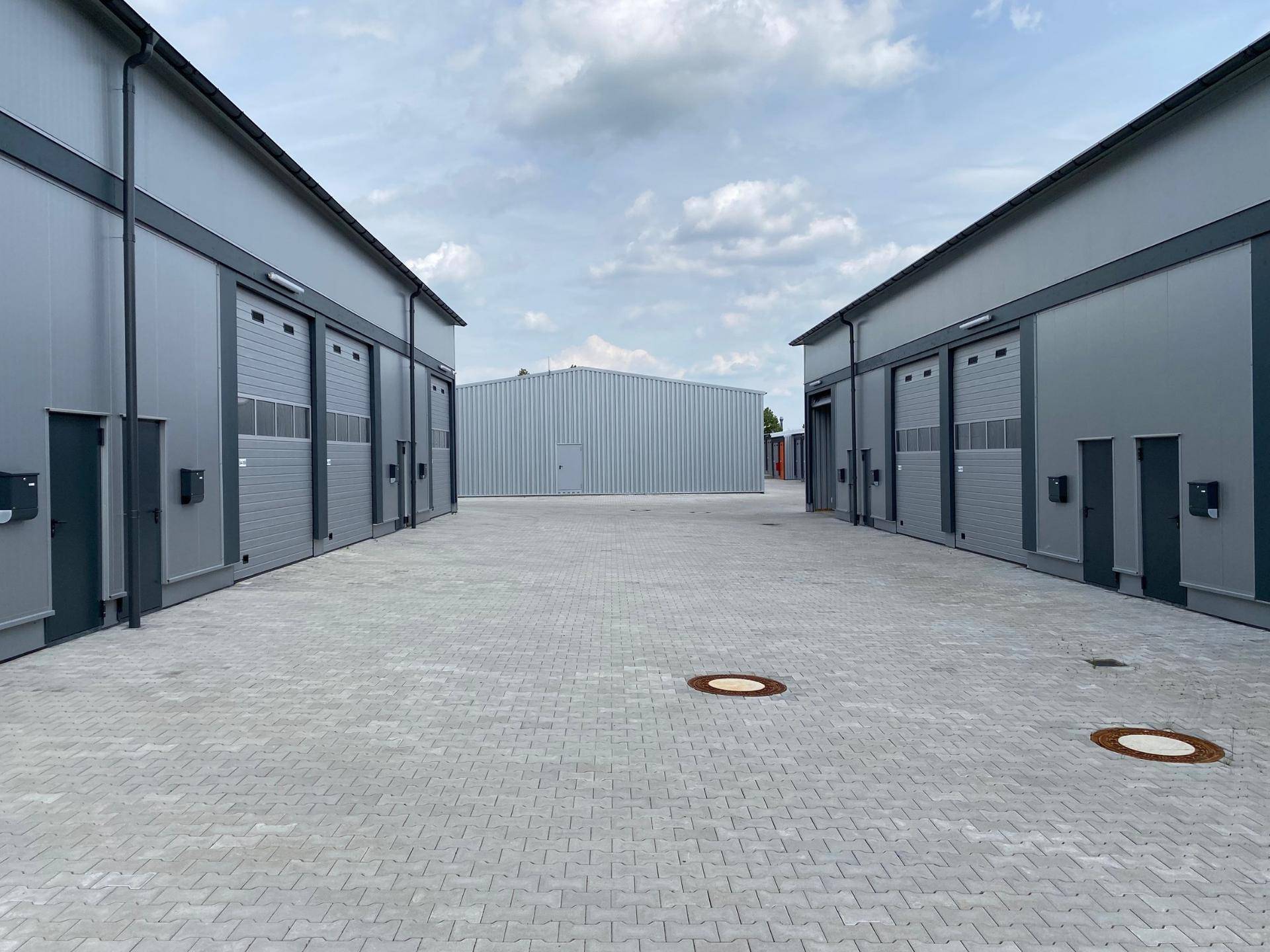 Industriekomplex mit zwei Reihen grauer Gebäude, Rolltoren und gepflasterten Zufahrten unter bewölktem Himmel.