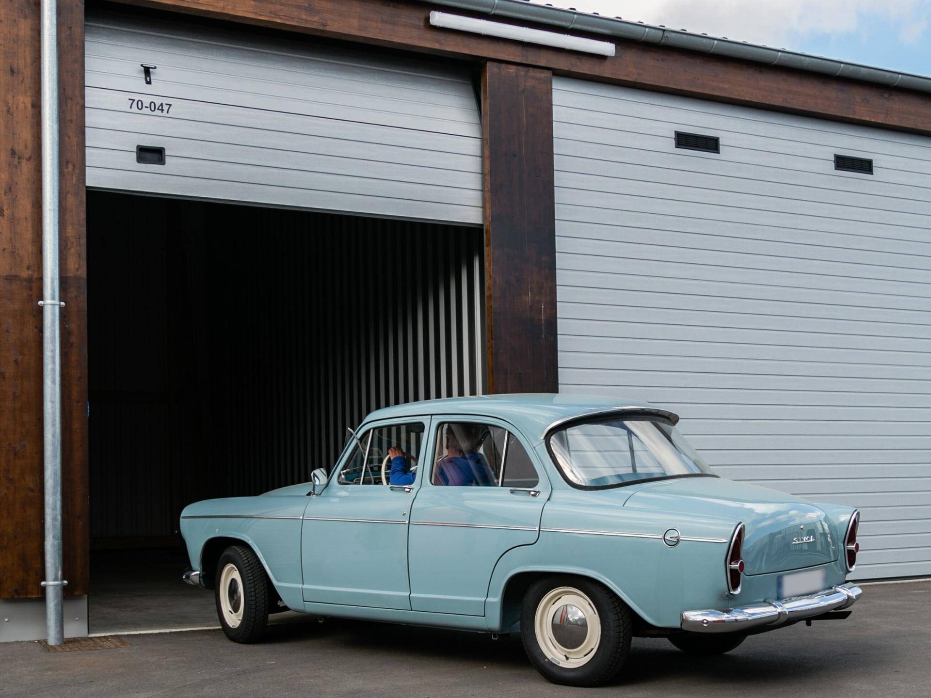 Ein hellblaues Oldtimer-Auto parkt teilweise in einer offenen Lagerhalle mit Holzelementen.
